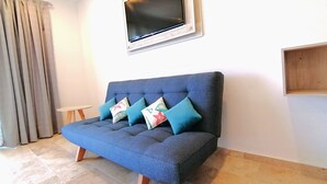 A blue sofa with four cushions, a mounted TV, and a small wooden shelf on a white wall.