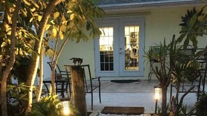 A pathway leading to a building with a door and windows, surrounded by plants and a small table. - Bougainvillea Cottage (Peggy's Retreat) (Hobe Sound)