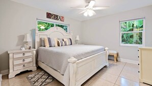 A bedroom with a bed, nightstands, a ceiling fan, a window with a view, and a small stool. - Bougainvillea Cottage (Peggy's Retreat) (Hobe Sound)