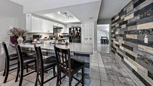 A modern kitchen with white cabinets, a large island with bar stools, a dining area with a table and chairs, and a decorative wall with a mix of wood and stone textures. - Bougainvillea Cottage (Peggy's Retreat) (Hobe Sound)