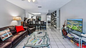 A modern living room with a leather sofa, a glass coffee table, a dining area with chairs, a kitchen in the background, and a large TV on the right. - Bougainvillea Cottage (Peggy's Retreat) (Hobe Sound)