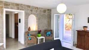 A living room with a stone wall, wicker chairs, a black sofa, a small table with a green pot, and a view through an open door. - Scrittore2 - Scrittore2 Villa Fiore (Capoliveri)