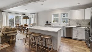 A modern kitchen with white cabinetry, a central island with stools, a dining area with a sofa and chairs, and a view of the outdoors through large windows. - Dog Friendly Resort Home at Topnotch Spa & Resort (Stowe)