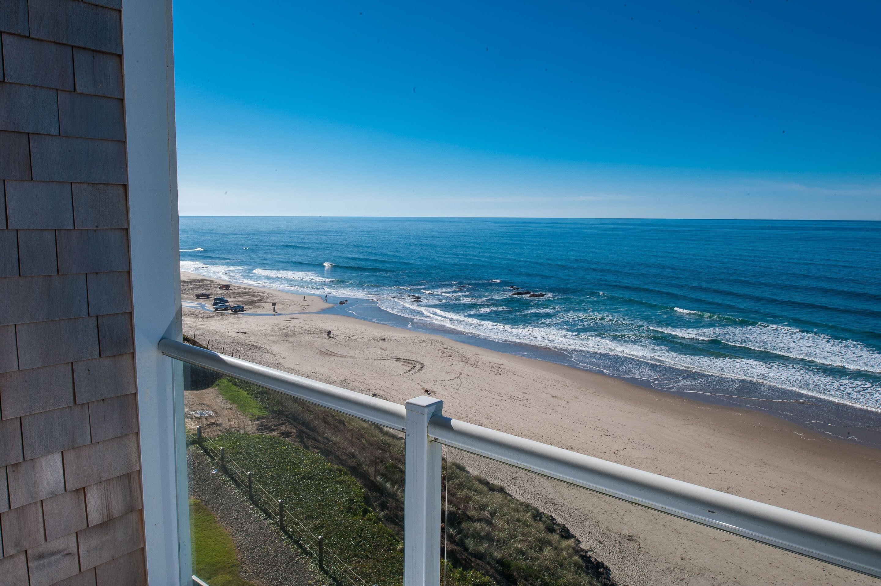 A beach view from a balcony with a railing, sandy beach, ocean waves, and a clear sky.