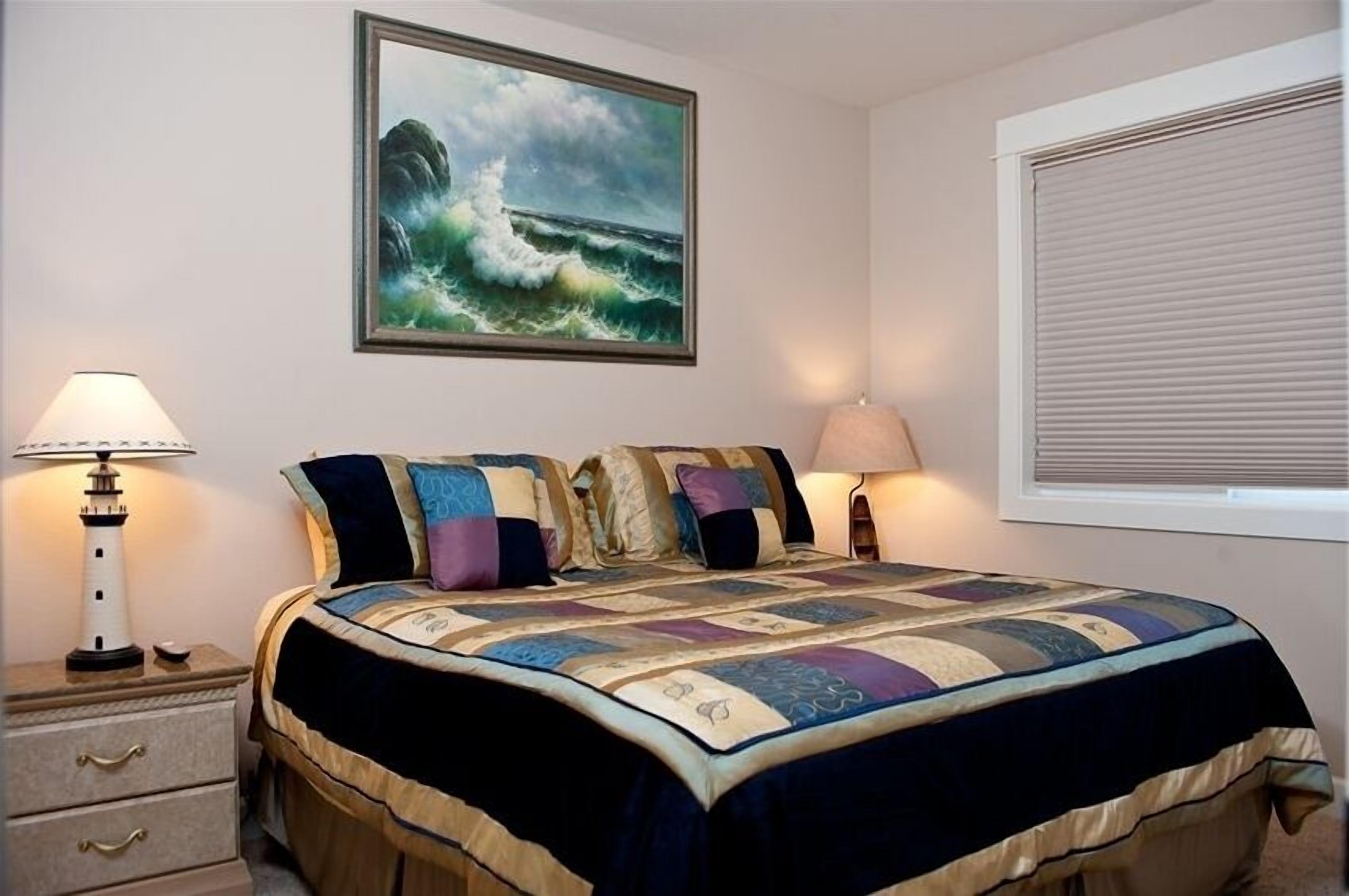 A bedroom with a large bed, two bedside lamps, a painting of a wave, and a window with blinds.