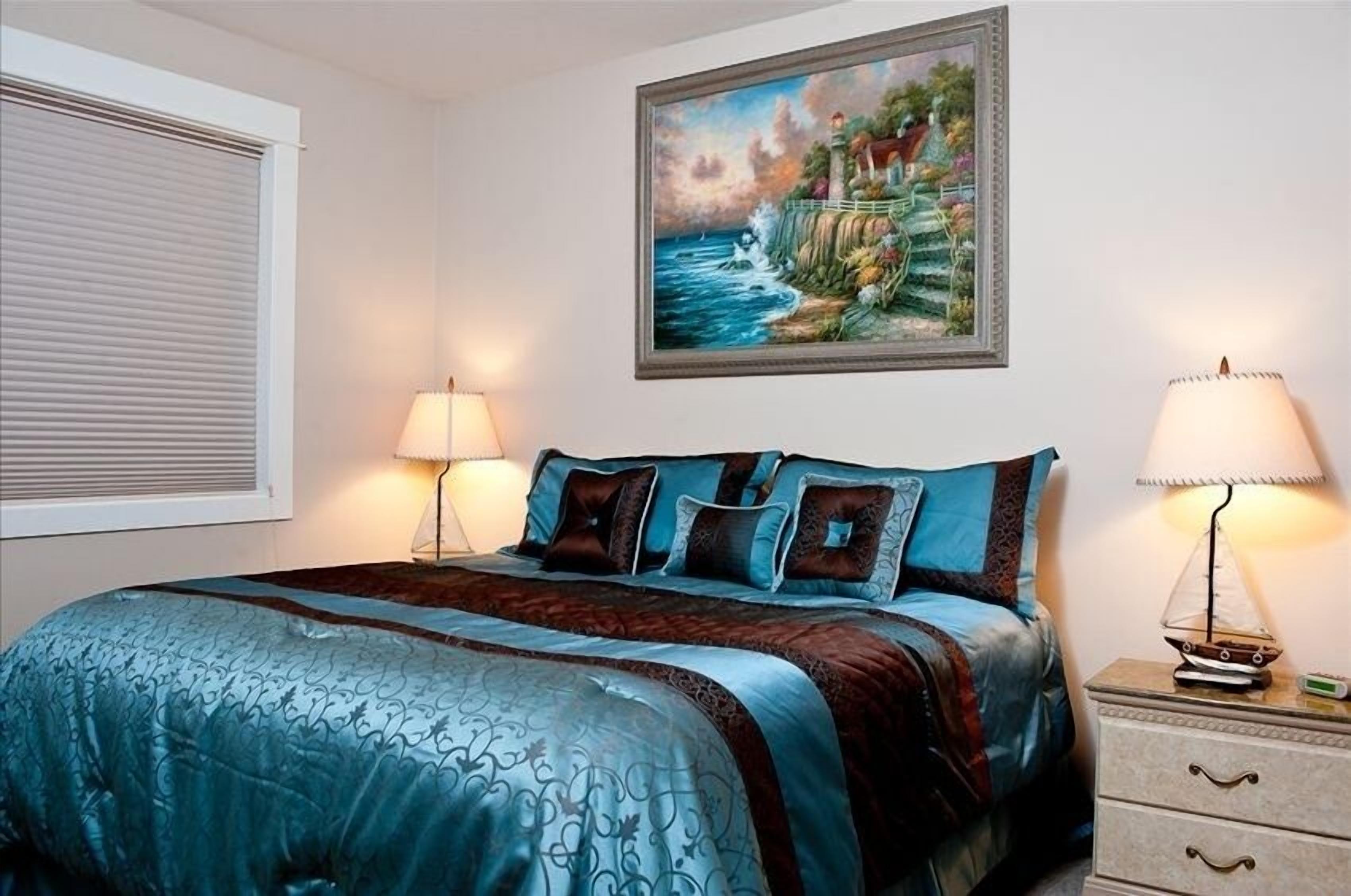 Un dormitorio con una cama grande, dos lámparas de noche, una mesita de noche con lámpara y un cuadro enmarcado en la pared.