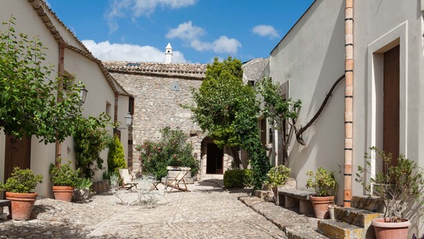 Une cour pavée de pierres, bordée de murs en pierre, agrémentée de plantes en pot et dotée d’un espace de repos central.