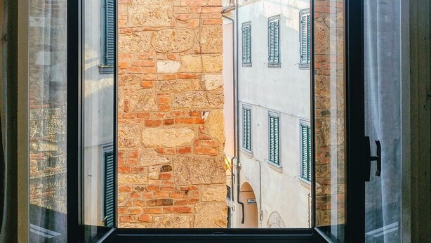 Une vue par une fenêtre dont les volets sont ouverts, dévoilant un bâtiment en pierre surmonté d’un dôme et coiffé d’un toit rouge.