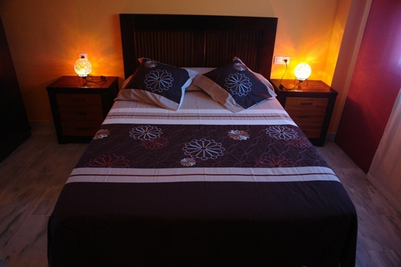 A neatly made bed with a patterned bedspread and two decorative pillows. There are two bedside tables with lamps, and the room has wooden flooring.