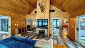A log cabin interior with a fireplace, a television, a sofa, and a deer head mounted on the wall. - Sandpiper Too | West Twin Lake Home (Lewiston)