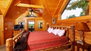 Dormitorio de cabaña de madera con cama, ventilador de techo, televisor y ventana con vista a follaje.