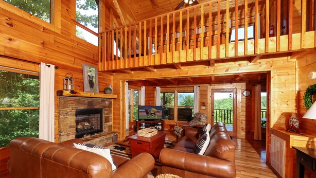 Interior de una cabaña de madera con chimenea, sofás de cuero, televisor y balcón.