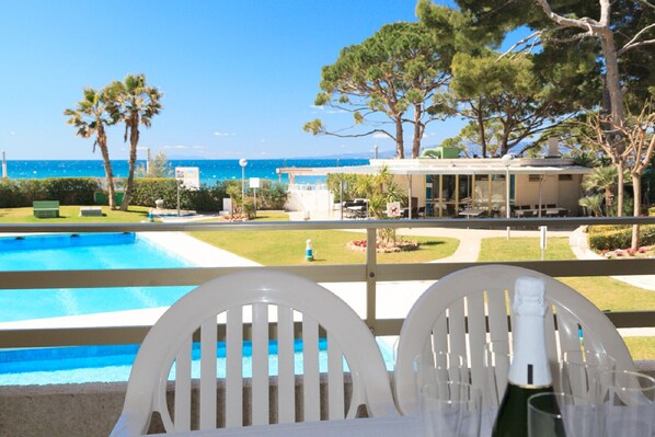 A poolside view with a clear view of the ocean, a covered outdoor area, and a building with a patio.