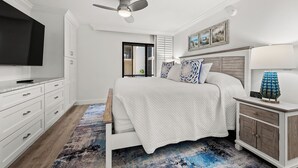 A bedroom with a bed, a TV, a ceiling fan, a nightstand, and a painting on the wall.