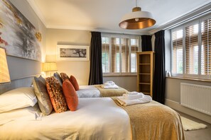 A hotel room with two beds, a large window with blinds, a hanging light fixture, and a painting on the wall.