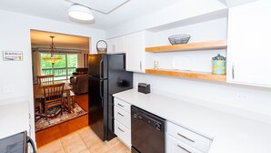 Une cuisine moderne avec des meubles blancs, un réfrigérateur noir et un lave-vaisselle. On y trouve un coin repas avec une table et des chaises, ainsi qu’une fenêtre donnant sur un paysage verdoyant.
