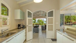 A modern kitchen with white cabinets, a stainless steel sink, and a coffee maker. There is a view of a patio area with outdoor furniture and a view of the ocean. - Areca Palm - luxury villa with private pool, sea and countryside views (Le Vauclin)