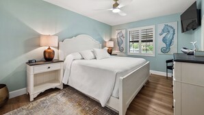 Quarto com cama, mesas de cabeceira, ventilador de teto, obras de arte e escrivaninha com cadeira.