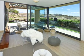A modern interior with a glass wall, a white furry chair, and a round grey ottoman. - Salobre Los Lagos 26 (Maspalomas)