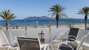 A beachside table with white chairs, a bottle of champagne, and a glass of wine, overlooking a beach with umbrellas and people.