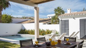 A poolside dining area with a table set for breakfast, overlooking a swimming pool and a white building with shutters.