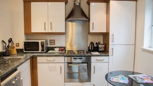 Una cucina compatta con mobili bianchi, una cappa in acciaio inossidabile, un forno a microonde, un bollitore e un piccolo tavolo da pranzo con riviste.