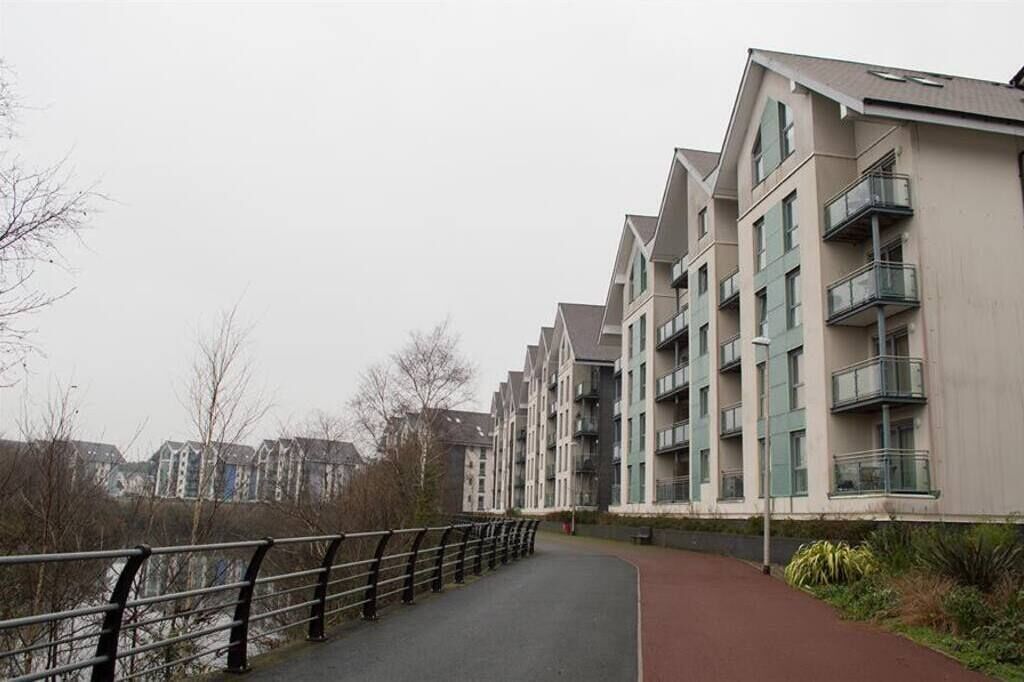 A modern apartment building with balconies, a paved walkway, and a river in the background.