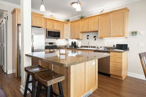 A kitchen with wooden cabinets, a stainless steel refrigerator, a microwave, a stove, and a sink. There are two bar stools and a clock on the wall. - Promo! Beautiful Oceanfront Condo with Private Hot Tub, Pool, Wifi and More! (Lincoln City)