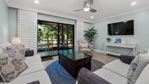 A modern living room with a glass coffee table, a ceiling fan, a TV, and a view of the outdoors. - Newly Renovated Garden View 2 bed, 2 bath at Sanibel Moorings #641 (Sanibel)