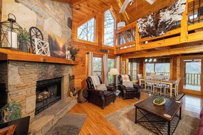 A spacious log cabin interior with a stone fireplace, leather seating, a dining area, and large windows offering views of the outdoors.