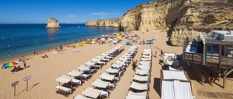 A beach with rows of sunbeds and umbrellas, a lifeguard tower, and a rocky cliff in the background.