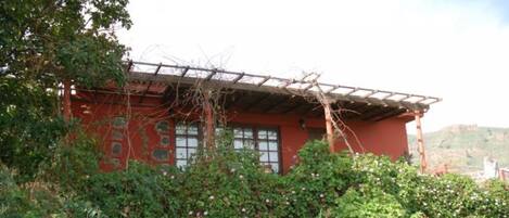Un edificio rojo con pérgola de madera, rodeado de abundante vegetación y plantas florecientes.