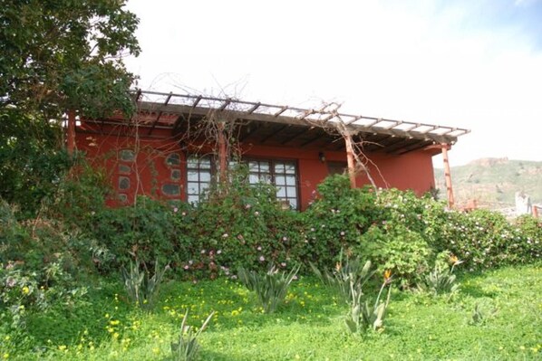 A red building with a wooden pergola, surrounded by lush greenery and flowering plants.