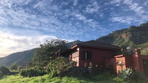 Una casa roja con un muro de piedra, rodeada de follaje y colinas.