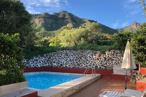 Piscina con muro de piedra y escalones, rodeada de vegetación y vista a la montaña.
