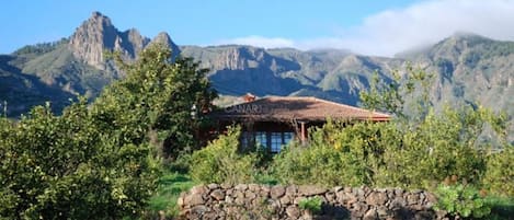 Un edificio rústico con techo de tejas, rodeado de vegetación y un muro de piedra, con montañas al fondo.