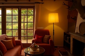 Ein Zimmer mit Sofa, Sessel, Tisch mit Obstschale, Lampe und Kamin, an dem ein Hirschkopf hängt.