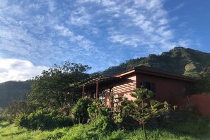 Une maison dotée d’un porche abrité, entourée d’une végétation luxuriante et ayant une chaîne de montagnes en toile de fond.