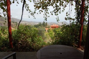 A view from a covered outdoor area with a wooden table, surrounded by greenery and a distant view of a red-roofed building.