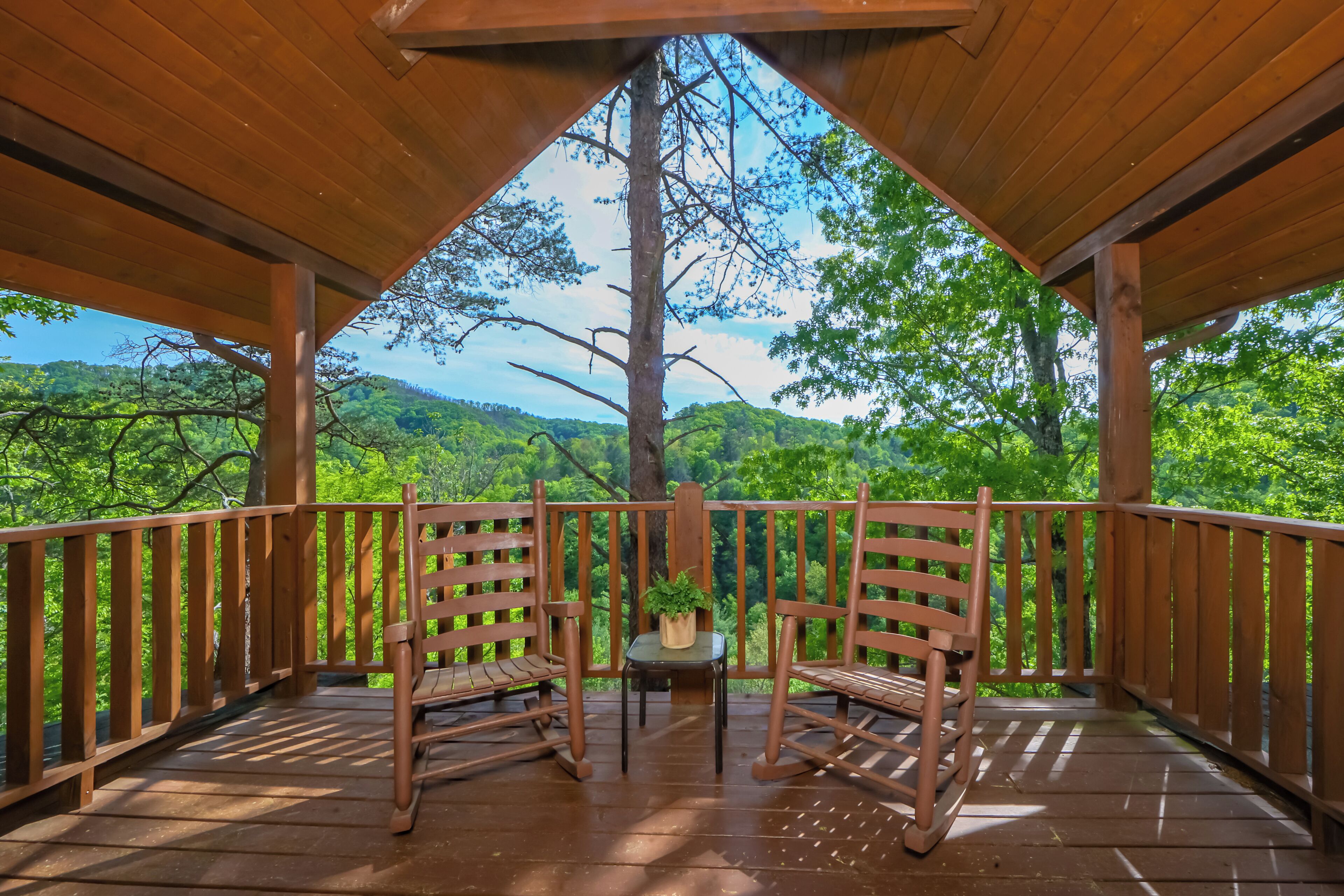 Una terraza de madera con dos mecedoras y una pequeña mesa, con vista a un paisaje verde exuberante.