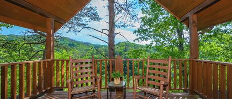 Una terraza de madera con dos mecedoras y una pequeña mesa, con vista a un paisaje verde exuberante.