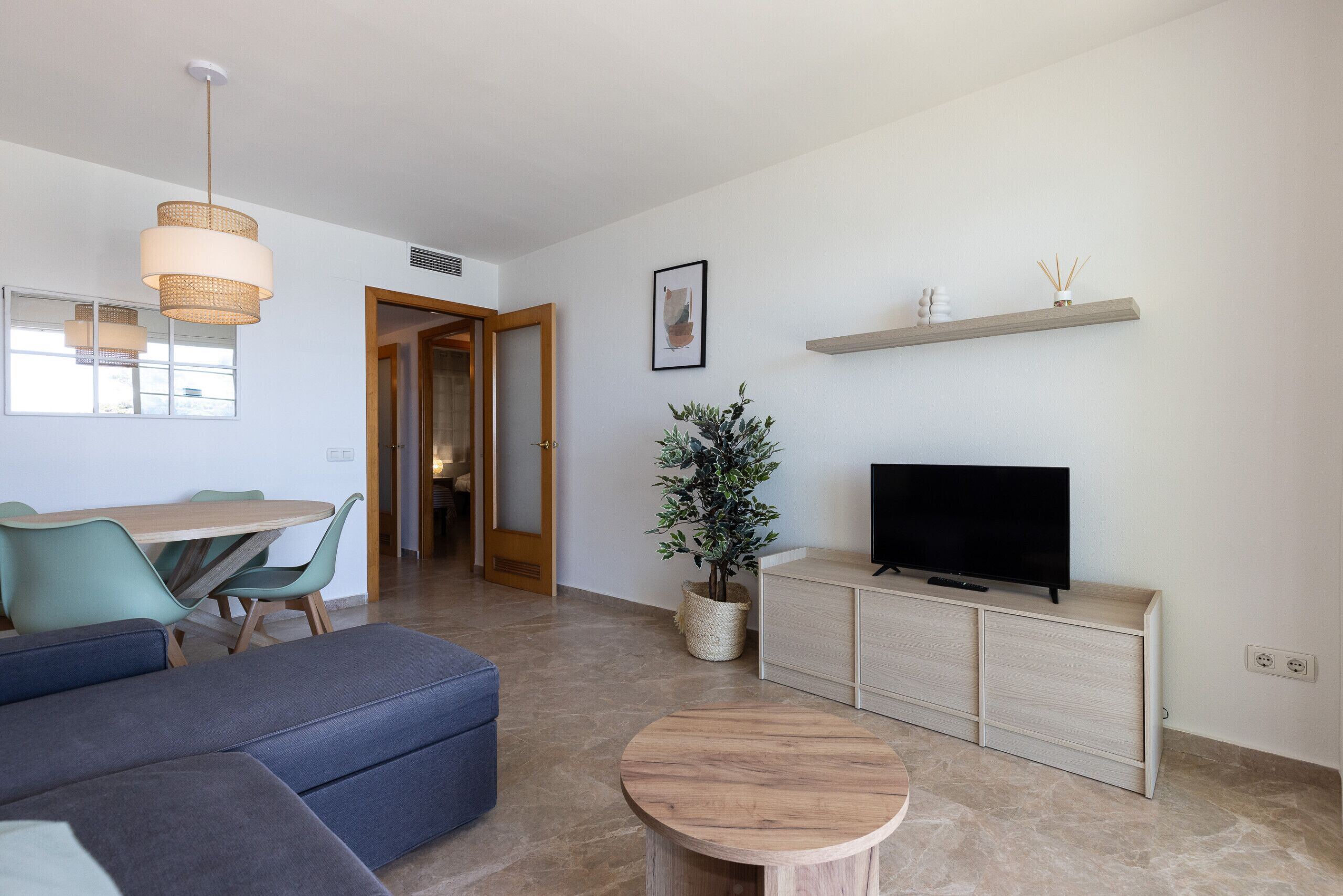 Una sala de estar moderna con un sofá, una mesa redonda de madera, un televisor sobre un mueble bajo, una planta en maceta y una zona de comedor con mesa y sillas.