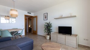 A modern living room with a sofa, a round wooden coffee table, a television on a cabinet, a potted plant, and a dining area with a table and chairs. - Family Holiday with spectacular sea views · UHC ARINSAL (Salou)