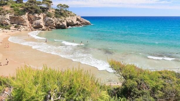 A beach with clear turquoise waters, rocky cliffs, and green vegetation. - Family Holiday with spectacular sea views · UHC ARINSAL (Salou)