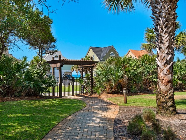 A pathway leading to a covered gazebo in a well-maintained garden with palm trees and a house in the background.