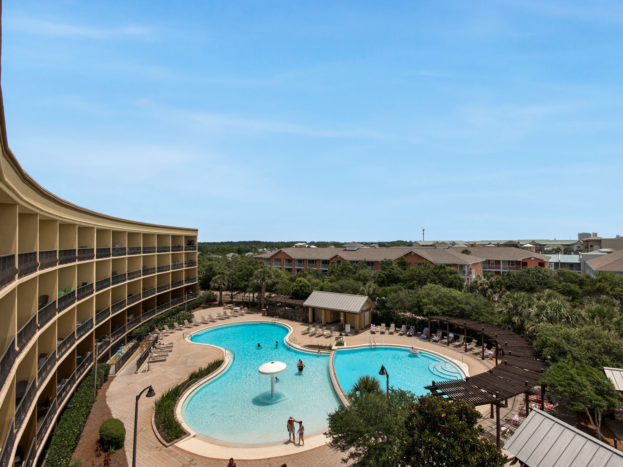 A hotel with a large, curved swimming pool surrounded by lounge areas and a multi-story building with balconies.
