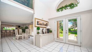 A spacious indoor area with a tiled floor, a central seating area with chairs and a table, and a door leading to an outdoor garden. - SWISS 41 - Golf Course (Clermont)