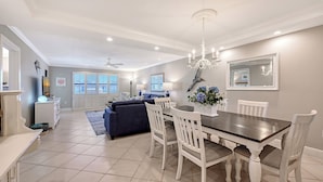 A spacious living area with a dining table, white chairs, a chandelier, and a large window with shutters.