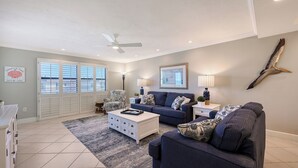 A spacious living room with a large window, a ceiling fan, and a decorative bird on the wall.