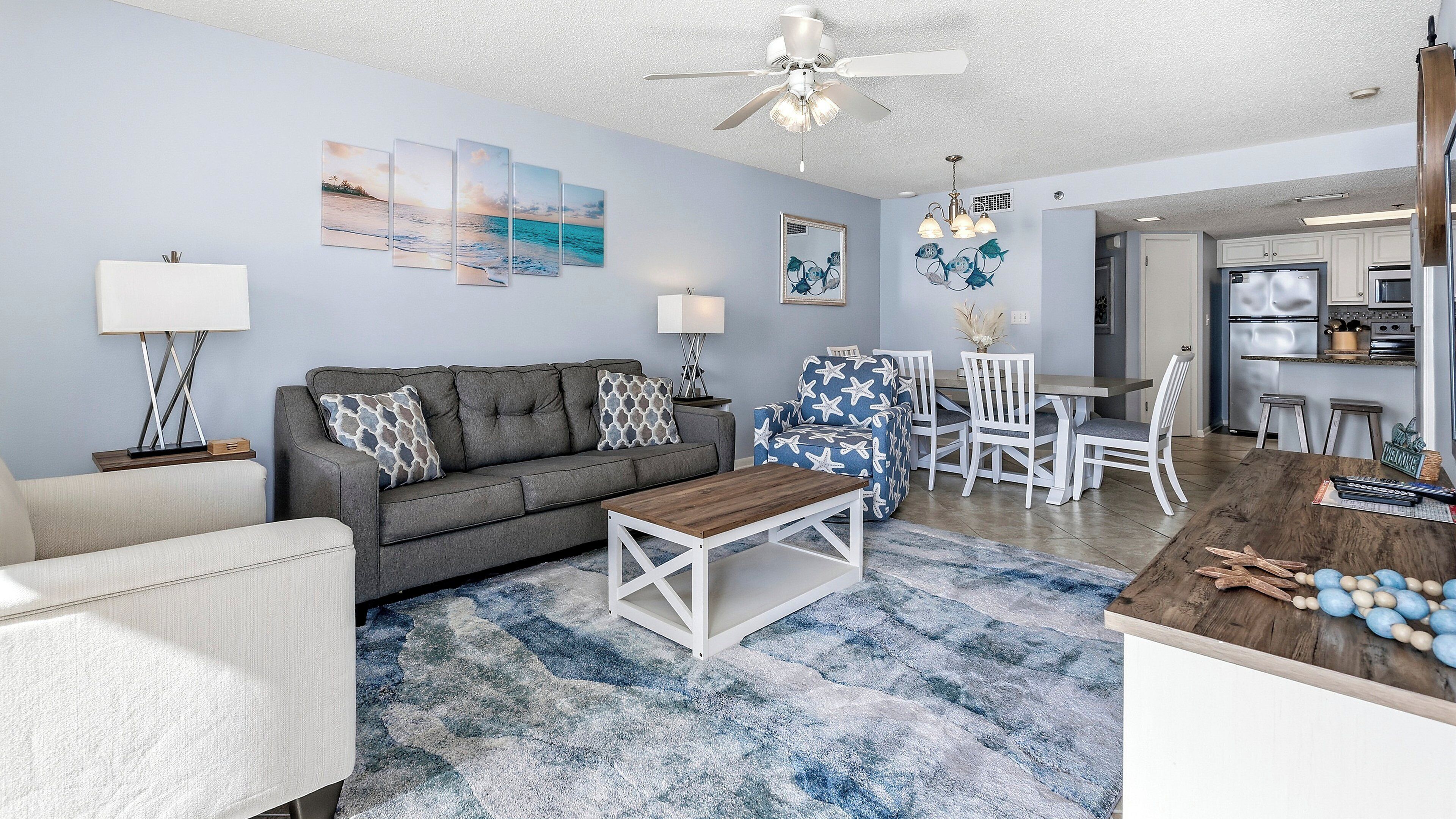 Ein Wohnzimmer mit grauem Sofa, einem Couchtisch und einem blau gemusterten Sessel. Im Hintergrund befindet sich eine Küche mit weißen Schränken und ein Essbereich mit Tisch und Stühlen.